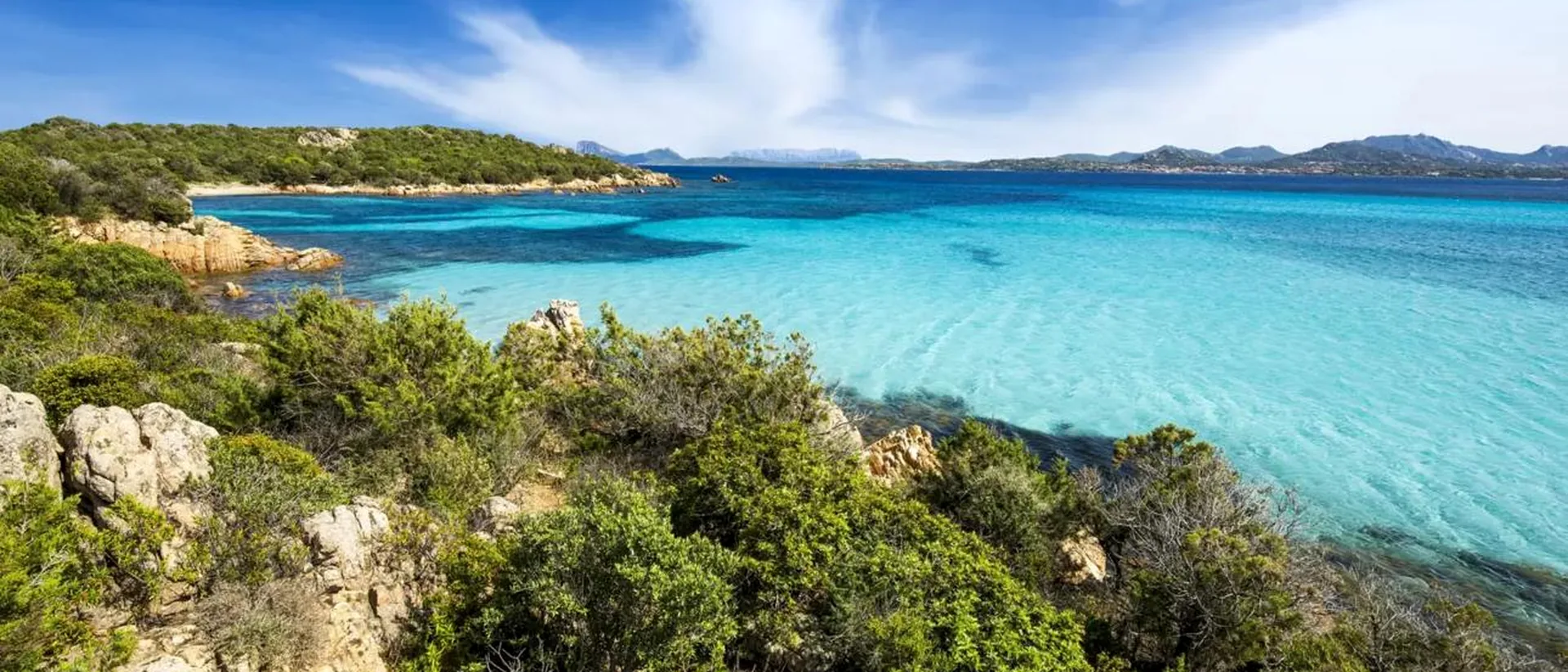 Rocky Sardinian shoreline, green Mediterranean vegetation and bright blue water.