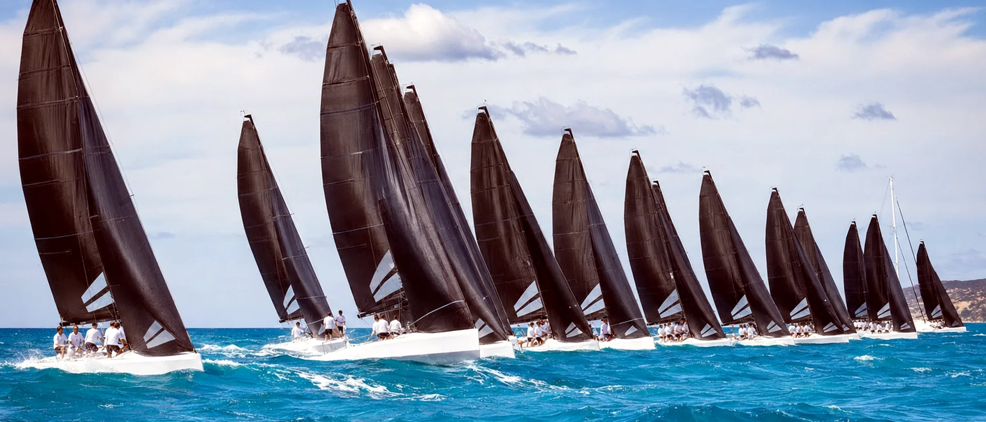 Fleet of racing sailboats with black sails crossing bright turquoise water.