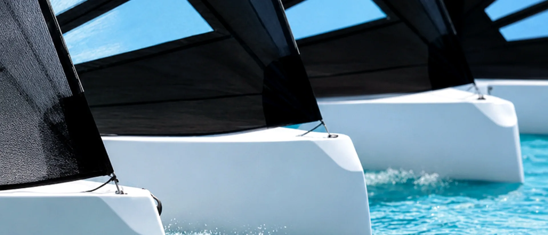Close-up line of white hulls and black sails moving over turquoise water.