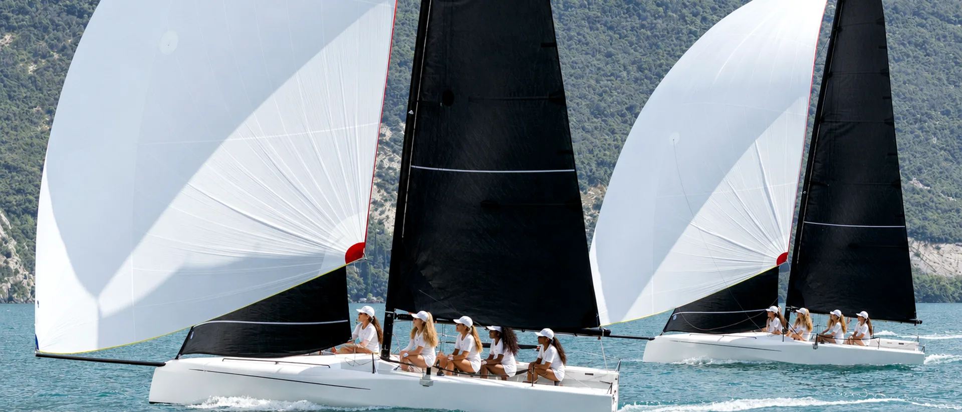 Two white racing sailboats with black mainsails and white spinnakers on mountain-backed water.