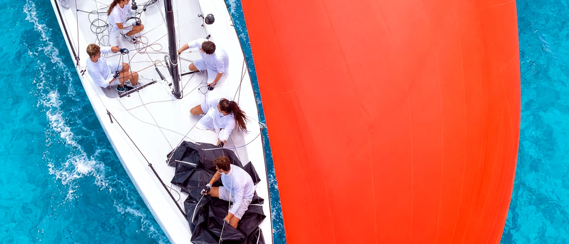 Aerial view of a white sailboat with a large orange spinnaker over clear turquoise water.