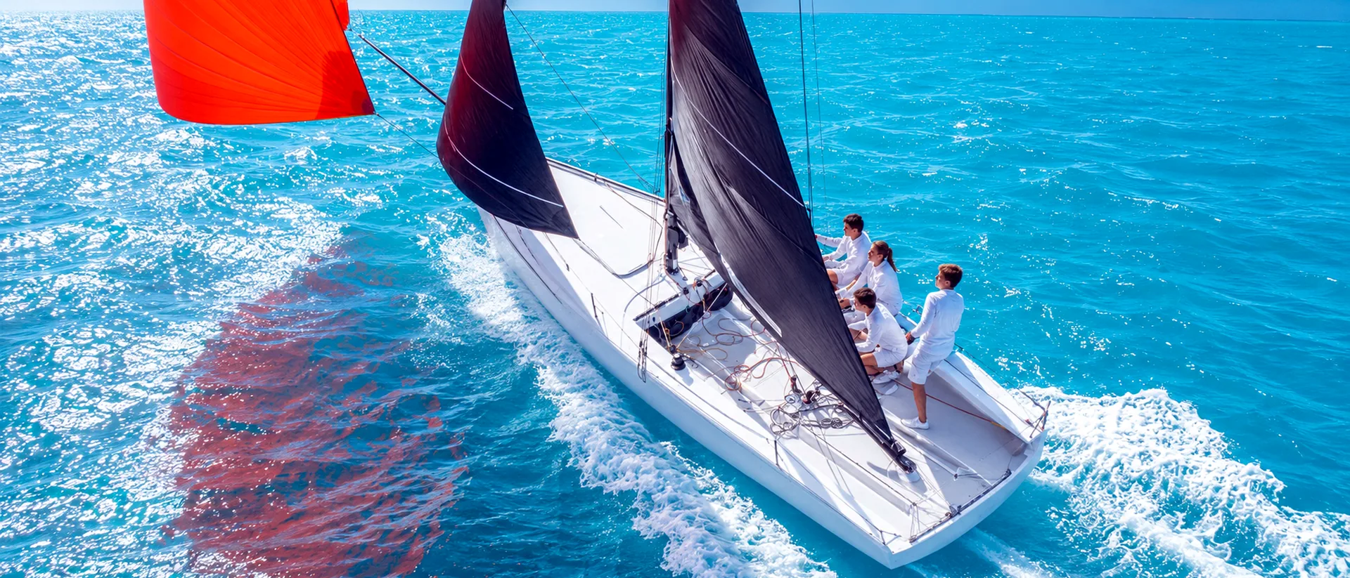 Aerial view of a white sailboat sailing fast through turquoise water with black sails and an orange spinnaker.