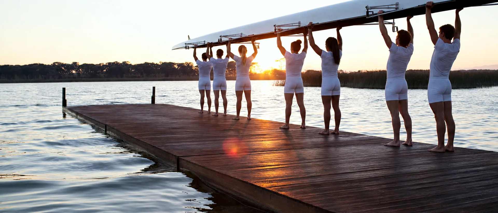 Rowing crew carrying a shell over a dock at sunrise.
